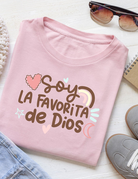 Folded pink t-shirt with 'Soy la Favorita de Dios' text on a white surface with sunglasses and shoes.