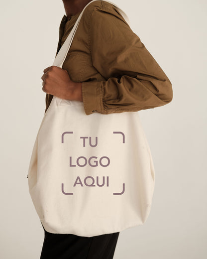 Beige tote bag with 'TU LOGO AQUI' text held by a person wearing a brown jacket.